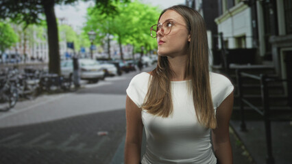 Fototapeta premium Young woman with glasses looking up while standing on a street beside parked bicycles and a building, wearing a white shirt and long hair; contemplation.