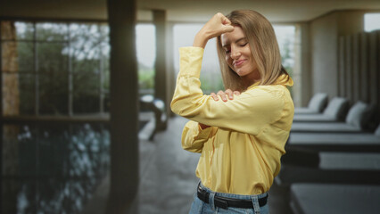 Fototapeta premium Woman flexes bicep with folded arm in yellow shirt at an indoor spa pool lounge inside a building; confidence empowerment.