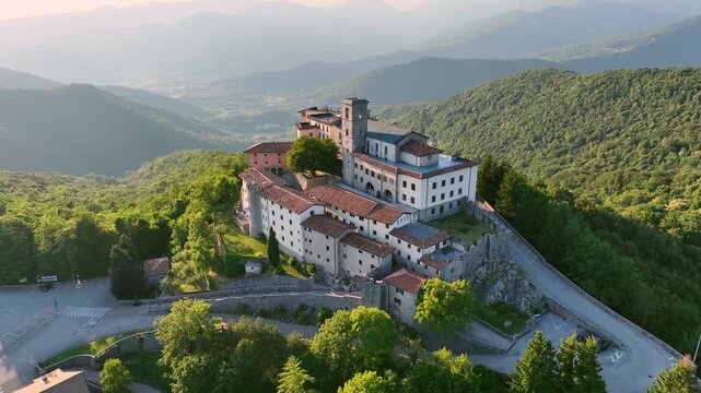 Majestic drone shot displays historic sanctuary of Castelmonte on hilltop. Ancient stone fortification walls and religious architecture stand over Friuli region landscape under clear sky.