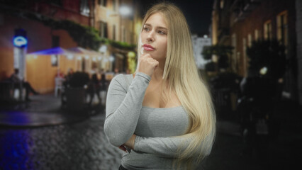 Fototapeta premium Young blonde woman with hand on chin and folded arm wearing a grey long sleeve top, standing by a cobblestone street cafe under lights at night; contemplation.