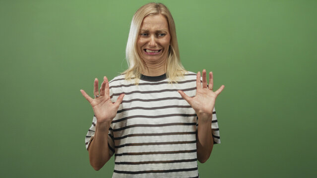 Woman middle aged blonde in striped tshirt, palms raised and fingers splayed with a grimace against a green studio backdrop; disgust reluctance.