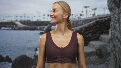 Fototapeta premium Woman smiles and tilts head, showing exposed midriff and maroon tank top in front of a seaside building with volcanic rocks; joy.