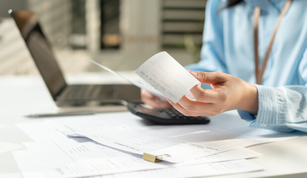Businesswoman hands review receipts beside laptop and calculator. Focused expression shows careful financial management. Desk cluttered with papers implies busy work environment.