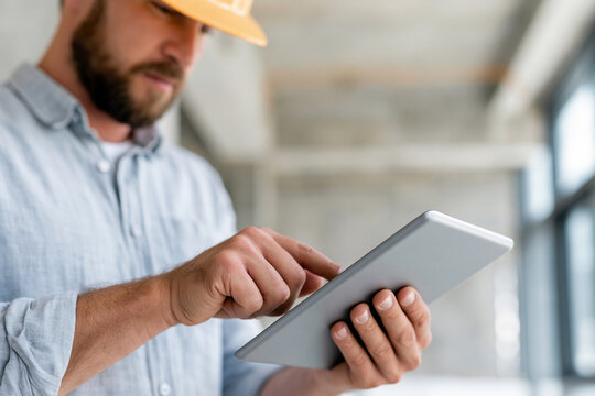 Bearded man wears a hard hat and checks a tablet. His focused gaze suggests planning or inspection. Concrete walls hint at a construction or renovation site. He holds the device with purpose