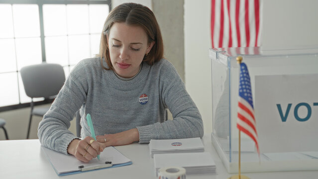 Woman writing on clipboard with pen at voting table beside clear ballot box and small us flags in a polling building; patriotic civic duty.