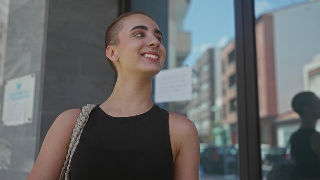 Woman with short hair smiling showing teeth, bare shoulder and braided bag strap, looking up on street; carefree joy.