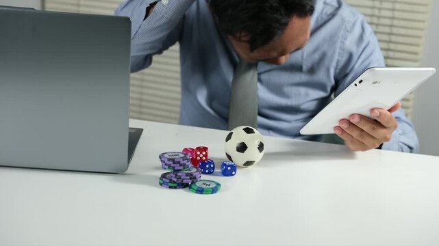 An Asian man holds a tablet and watches his favorite team lose. He feels sad because he lost a soccer bet, staring at the screen with a stressed expression. Gambling and soccer betting
