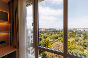 Fototapeta premium Hotel room window overlooking urban park, distant skyline, and clear blue sky with soft warm interior lighting, framed by sheer curtains, glass reflections, and spacious high floor view