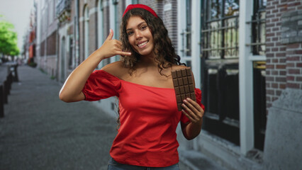 Obraz premium Woman in red shirt holding a chocolate bar and making a phone hand gesture in street by brick building windows; playful summer treat.