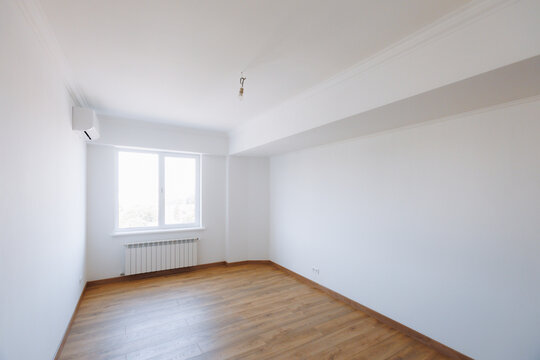 Bright empty room with wood floor, wall mounted air conditioner, radiator, window daylight, white walls, ceiling molding, hanging light bulb, and clean minimal residential interior