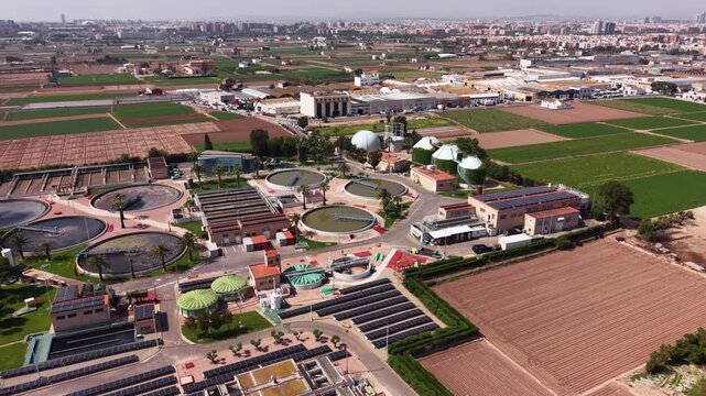 Aerial view of a wastewater treatment facility featuring circular clarifier tanks, solar panels, and anaerobic digesters surrounded by agricultural fields with a city skyline in the distance
