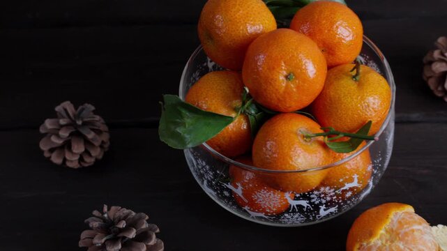 Glass Christmas bowl filled with fresh tangerines on a dark wooden table. Peeled mandarin and pine cones create a cozy winter holiday atmosphere. Festive seasonal food background