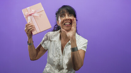 Fototapeta premium Senior hispanic woman holds pink gift box with ribbon, lifting it and pressing hand to cheek in a surprised gesture in studio with purple backdrop; surprise celebration.