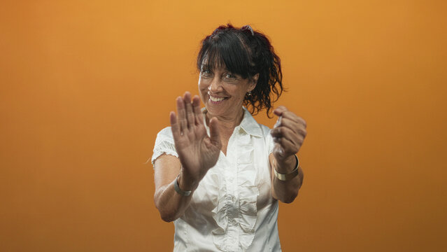 Woman senior smiling miming steering wheel with raised palm and outstretched hands in studio orange wall wearing white blouse; joy warmth.