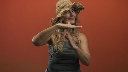 Fototapeta premium Woman makes time out gesture with palms horizontal and visible hands near chest, wearing straw hat and sleeveless top against orange backdrop in studio; confidence pause.