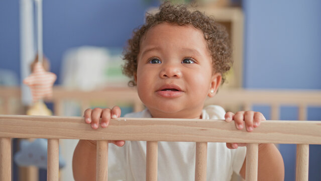 Little girl stands and grips crib rail with small hands in nursery building near mobile; tender curiosity warmth.