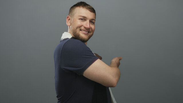 Man with soccer scarf pointing with bare forearm and smiling while turning in gray studio set; cheerful mischief.
