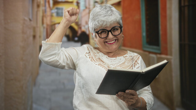 Woman plus size reader with grey hair and glasses reading a book, right fist raised and smiling while seated in a narrow european street; joy leisure empowerment.