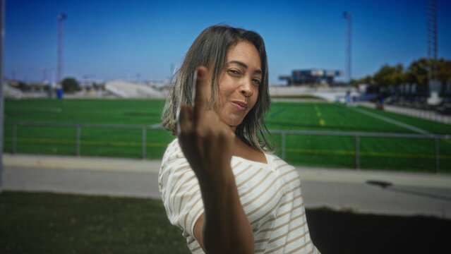 Hispanic woman showing middle finger to camera on a sunny athletic field with bleachers and fencing building; defiance.