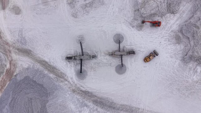 Overhead view of two mobile screening machines with conveyor arms creating circular stockpiles on pale gypsum quarry surface, with excavator and wheel loader nearby