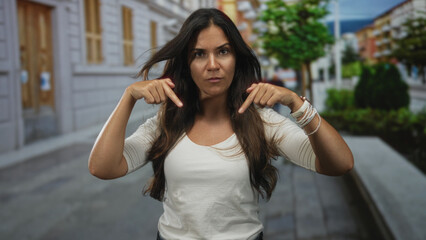 Fototapeta premium Young hispanic woman pointing both index fingers to chest on city street with trees and buildings blurred; confidence.