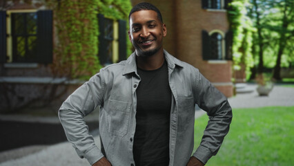 Fototapeta premium Black man smiles with hands behind back, open grey shirt over black tee, visible face and relaxed tilt in front of a brick building; confidence ease.