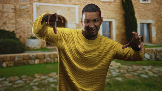 Young man makes claw gesture with both hands while wearing a yellow sweater in front of a stone building, playful grin visible; playfulness energy.