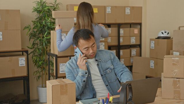 Man talking on smartphone at desk and gesturing with hand while woman lifts and stacks cardboard packages on shelves in building; small business productivity.