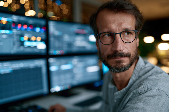 Man with glasses stares at multiple screens. He appears focused, slightly tired, and thoughtful. Behind him, city lights blur softly in background