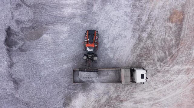 Aerial top view of a wheel loader dumping gypsum material into a truck trailer with dust rising during loading at an open pit quarry mining site