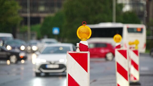 Row of reflective construction warning barrier posts with active orange yellow safety warning beacons on a wet road surface in the rain