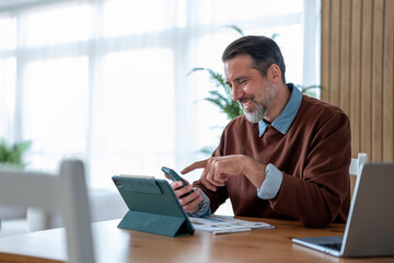 Fototapeta premium Man smiling using smartphone multitasking at home office