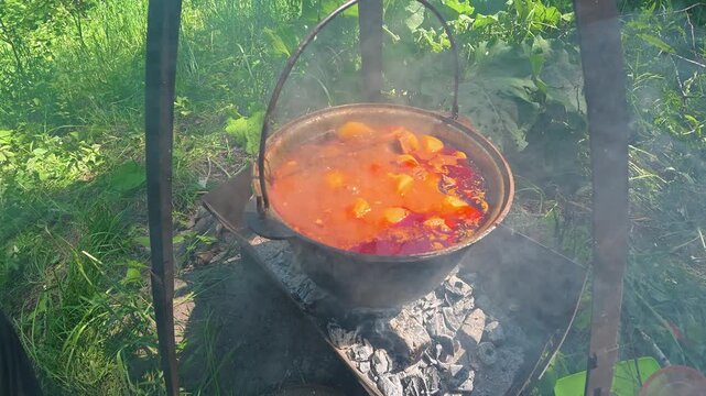 Red goulash soup boiling in cast iron cauldron over open campfire flame outdoors