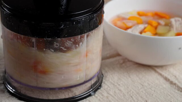 A blender starts processing a light chicken vegetable soup into puree while a bowl with the same diet broth stands nearby. Healthy homemade meal preparation