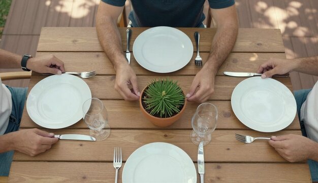 Wooden dining table with white plates, cutlery, glasses, and potted plant centerpiece, calm pre-meal atmosphere, outdoor terrace setting with some persons in frame