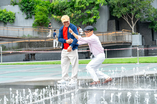 Couple Asian senior lgbtq men enjoy to play with fountain in the public park together and happiness during relax in the big city.