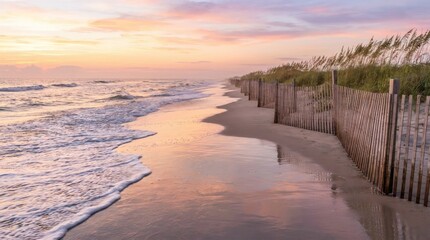 Fototapeta premium Sunrise on a tranquil beach with rolling ocean waves, sandy shore, protective dune fence, and colorful sky reflections.