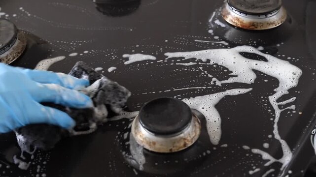  Cleaning black metal gas stove surface. Hand in rubber glove scrubs burner area with sponge and foam.