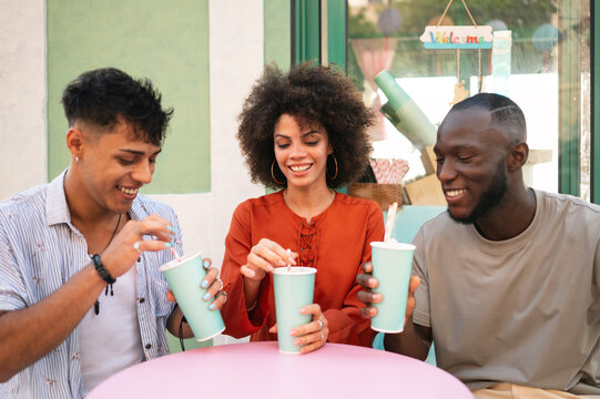 Diverse young friends enjoying contemporary cafe culture, laughing, toasting outside on a summer day