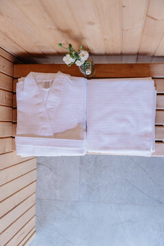 Vertical white bathrobe and folded towels with flowers arranged on wooden sauna bench