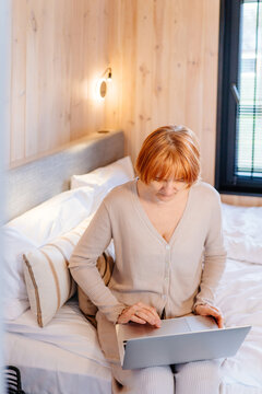 Vertical mature red-haired woman working remotely on laptop while sitting on bed in cozy cabin