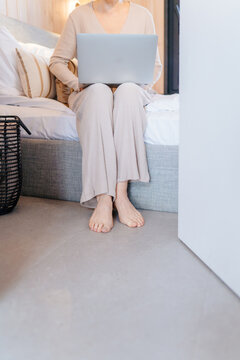 Vertical cropped woman using laptop while sitting barefoot on bed in modern cabin bedroom