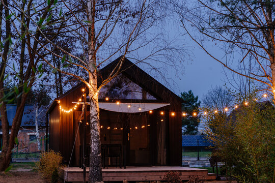 Cozy modern tiny cabin illuminated with string lights among trees during peaceful blue hour