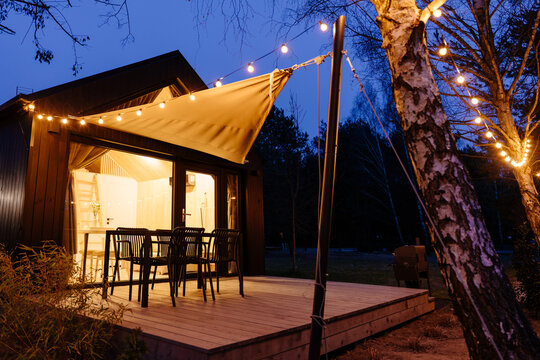 Modern tiny cabin terrace with dining furniture, canopy and string lights at blue hour