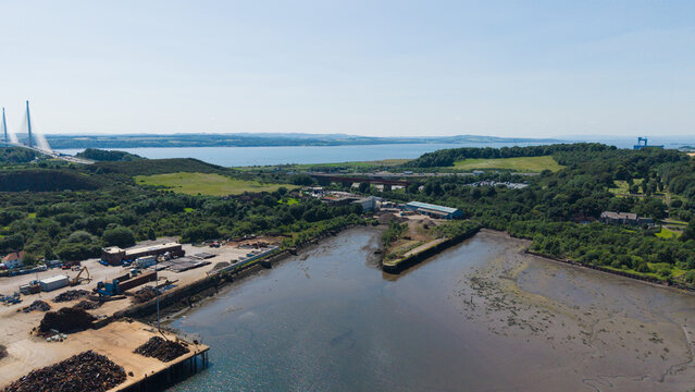 Aerial drone view of the coastline near Prestonhill Quarry in Inverkeithing, Fife, featuring the modern Queensferry Crossing bridge towers on the horizon under a clear sky in Scotland