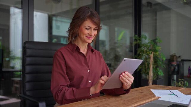 Middle aged businesswoman using digital tablet in office with copy space. Smiling mature woman financial advisor holds device, checks business data online. Female expert consults clients at desk.
