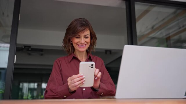 Using smartphone at office desk, older mature businesswoman texts client message. Smiling woman sales manager holds mobile phone. Female seller checks business app and reads offer at workplace.