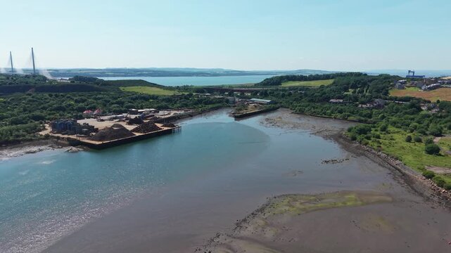 Aerial forward flight over Inverkeithing bay in Fife, Scotland, showing a muddy coast during low tide, industrial scrap yard pier, and distant Queensferry Crossing bridge under clear afternoon sun