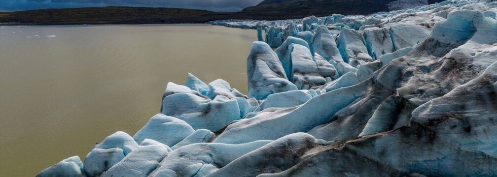 Panoramic view of Fjalls&aacute;rl&oacute;n Glacier Lagoon in southeast Iceland, where the majestic Vatnaj&ouml;kull Glacier meets calm waters beneath dramatic clouds and rugged volcanic mountains.