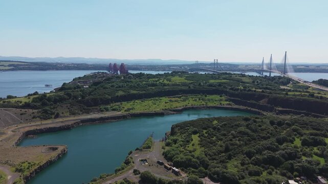 Aerial view of the deep blue lake inside Prestonhill Quarry in Inverkeithing, Fife, Scotland, showcasing the industrial site surrounded by green fields with three Forth bridges on the horizon